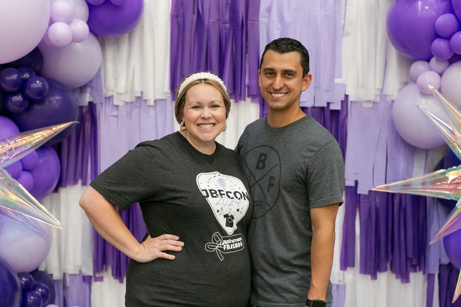 A mom with a large JBF shopping bag on her shoulder stands beside her husband who wears their toddler at a JBF sale.